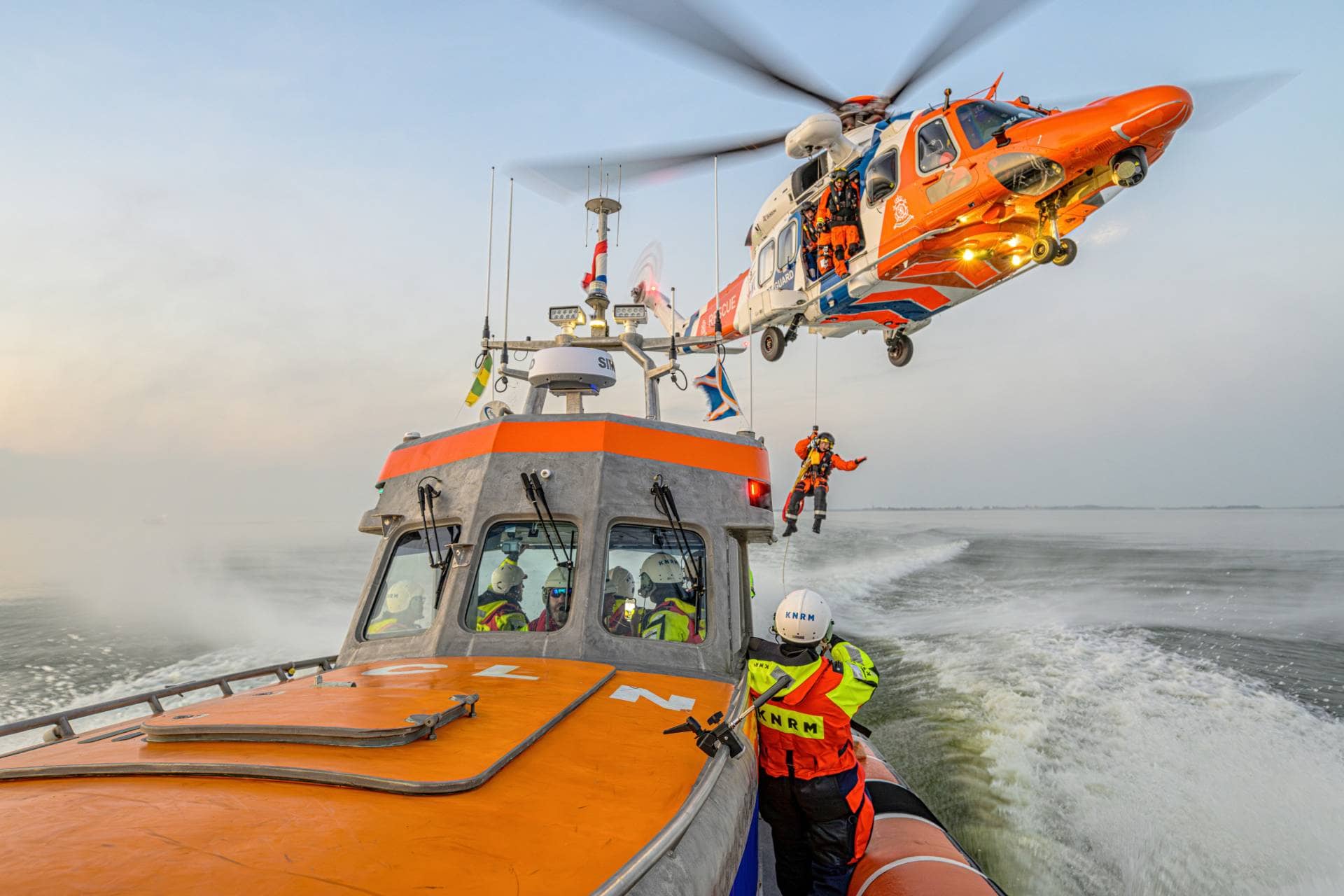 KNRM rescue vessel at sea with a rescue helicopter hoisting a crew member overhead